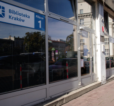 Na fotografii widać front budynku, przeszklone drzwi i duże witryny, po lewej i prawej stronie od wejścia. Na szybie tabliczka z napisem: „Biblioteka Kraków. Filia nr 1”. Przed wejściem stopień, przed budynkiem chodnik.