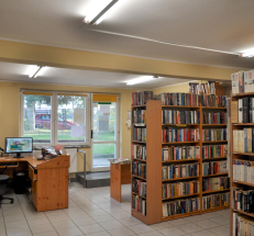 Na zdjęciu po lewej stronie lada biblioteczna ze stanowiskiem komputerowym, obok stół z czasopismami. Na wprost wyjście i jeden stopień z poręczą. Po prawej stronie drewniane regały z literaturą dla dorosłych czytelników. 