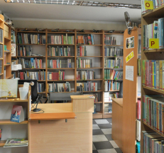 Na zdjęciu lada biblioteczna ze stanowiskiem komputerowym. W głębi pomieszczenia oraz z prawej strony widoczne regały z książkami. Na podłodze posadzka w kratkę. 
