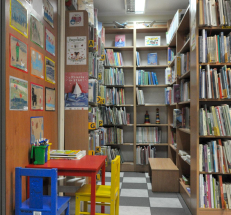 Na zdjęciu po lewej stronie drewniany stolik i krzesełka, nad nimi prace plastyczne dzieci, w głębi i po prawej stronie regały z literaturą dla młodszych czytelników, ławeczka.