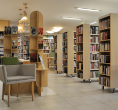 Na fotografii dział z literaturą dla dorosłych. Po lewej stronie lada biblioteczna, regały i stojaki na książki, fotel dla czytelników. Po prawej stronie regały w kolorze drewna z książkami. Na podłodze szara posadzka.