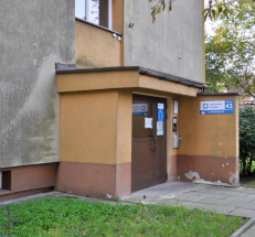 Na zdjęciu wejście do Filii. Drzwi wejściowe są szerokie, przed budynkiem chodnik z płyt chodnikowych. Na drzwiach tabliczki z godzinami otwarcia, na ścianie oznaczenie Biblioteki Kraków.