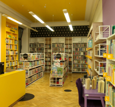 Na zdjęciu wzdłuż ścian żółte, białe i beżowe regały z literaturą dla dzieci i młodzieży, na środku niskie regały z książkami dla najmłodszych. Po prawej stronie stolik i krzesła. Ściany są kolorowe, na podłodze parkiet.