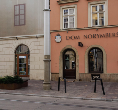 Na zdjęciu front budynku, w którym znajduje się Filia nr 2, oraz sąsiednią kamienicę (Dom Norymberski). Przed wejściem chodnik z kostką brukową, dwa niskie stopnie. Wejście jest przeszklone, na drzwiach tabliczka z godzinami otwarcia. 