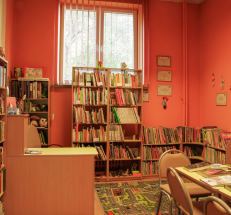 Na zdjęciu pomieszczenie z literaturą dla dzieci i młodzieży. Pod ścianami stoją wysokie i niskie regały z książkami, biurko, na środku stół i krzesła, naprzeciwko wejścia duże okno. Ściany mają kolor pomarańczowy, na podłodze kolorowy dywan.