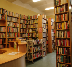 Na zdjęciu po lewej stronie stanowisko biblioteczne, obok niego wózek z książkami, wzdłuż ścian oraz w poprzek ustawiono regały z książkami. 