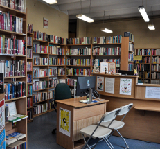 Na zdjęciu widoczne regały z książkami dla dorosłych ustawione wzdłuż ścian, z prawej strony lada biblioteczna, na niej monitor, figurki sów, plakaty. Na środku stoi nieduże stanowisko komputerowe z krzesłami.