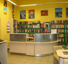 Na zdjęciu po prawej stronie stół i dwa fotele, po lewej stanowisko komputerowe z dostępem do katalogu elektronicznego, w głębi przejście do kolejnego pomieszczenia. Na wprost lada biblioteczna, za nią regały z książkami.