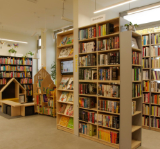Zdjęcie przedstawia po lewej stronie kącik dla dzieci: siedziska i regały w kształcie domków, dywan, z prawej strony wzdłuż ściany regały z książkami dla dzieci.