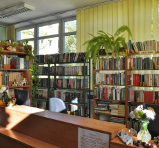 Na zdjęciu lada biblioteczna z dwoma stanowiskami, w głębi regały z książkami i duże okna częściowo przesłonięte roletami.