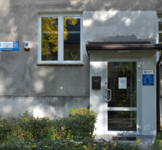 Na fotografii wejście do filii. Drzwi są przeszklone, na nich tabliczka z godzinami otwarcia, na ścianie obok oznaczenie Biblioteki Kraków. Do drzwi prowadzi brukowana alejka. 
