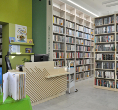 Na zdjęciu lada biblioteczna ze stanowiskiem komputerowym w kolorze jasnego drewna oraz regały z literaturą dla dorosłych w tym samym kolorze. Za ladą widać przejście do kolejnego pomieszczenia. 