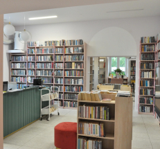  Po lewej stronie lada biblioteczna, na środku pomieszczenia oraz przy ścianach ustawione regały z książkami. Na ścianie ekran, nad ladą klimatyzator.