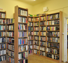 Na zdjęciu regały w kolorze drewna z książkami. Po prawej stronie kolejne pomieszczenie, w głębi widać stanowisko komputerowe, drukarkę.