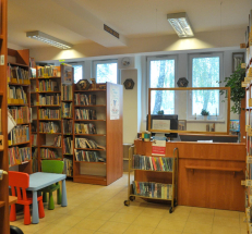 Na zdjęciu po lewej stronie regały w kolorze drewna z książkami oraz stolik z krzesełkami dla dzieci. Regały widać również w głębi pomieszczenia oraz po prawej stronie. Na wprost wózek z książkami, lada biblioteczna, za nią okno.