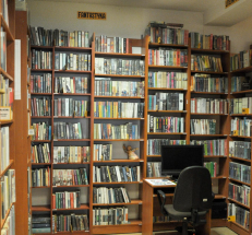 Na zdjęciu wzdłuż ścian regały w kolorze drewna, na nich książki dla dorosłych, pojedyncze stanowisko komputerowe dla czytelników.