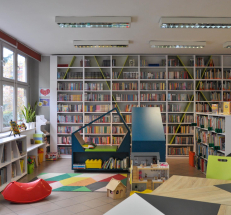 Na zdjęciu kącik dla dzieci. Na środku kolorowy dywan, siedziska w kształcie domków z półkami, domek dla lalek. Z lewej strony duże okna, pod oknami i z prawej strony nieduże regały, wzdłuż ściany wysokie regały z książkami.