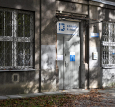 Na zdjęciu wejście do filii - budynek w szarym kolorze, przed budynkiem chodnik. Drzwi wejściowe są szerokie, w szarym kolorze. Po ich prawej i lewej stronie zakratowane, duże okna. Nad drzwiami oznaczenie Biblioteki Kraków.