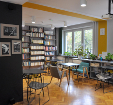 Na fotografii stoliki i krzesła dla czytelników, wzdłuż ścian ustawione regały z książkami. Z prawej strony duże okna, na parapetach rośliny.