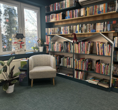 Po lewej stronie duże okna, przez które wpada światło, po prawej strony regały z książkami dla najmłodszych w kształcie domków, powyżej półki z książkami dla młodzieży. W kącie fotel, na podłodze dywan.