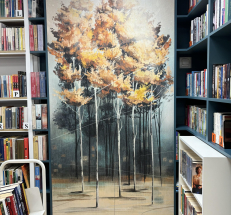 Na zdjęciu po prawej i lewej stronie drewniane regały z książkami w kolorze białym i szaroniebieskim, na środku sięgający sufitu panel z grafiką przedstawiającą brzozy.